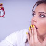 A surprised woman with yellow nails holds a small red alarm clock against a white backdrop.